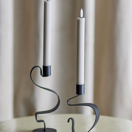 Ponza LED Candles Pack of 2 in beige, tall ribbed paraffin-look columns with flame-shaped tips, shown in black curved metal holders against a neutral curtain backdrop.