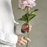 Person in white shirt holding the Peony Artificial Stem - Pink, showing its full ruffled bloom and leafy green stem against a neutral background.