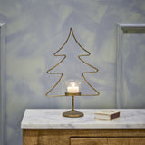 Miki Iron Christmas Tealight Holder in gold, a wire fir-tree outline with glass tealight cup and metal foot, shown lit on a marble-top sideboard beside a small book.