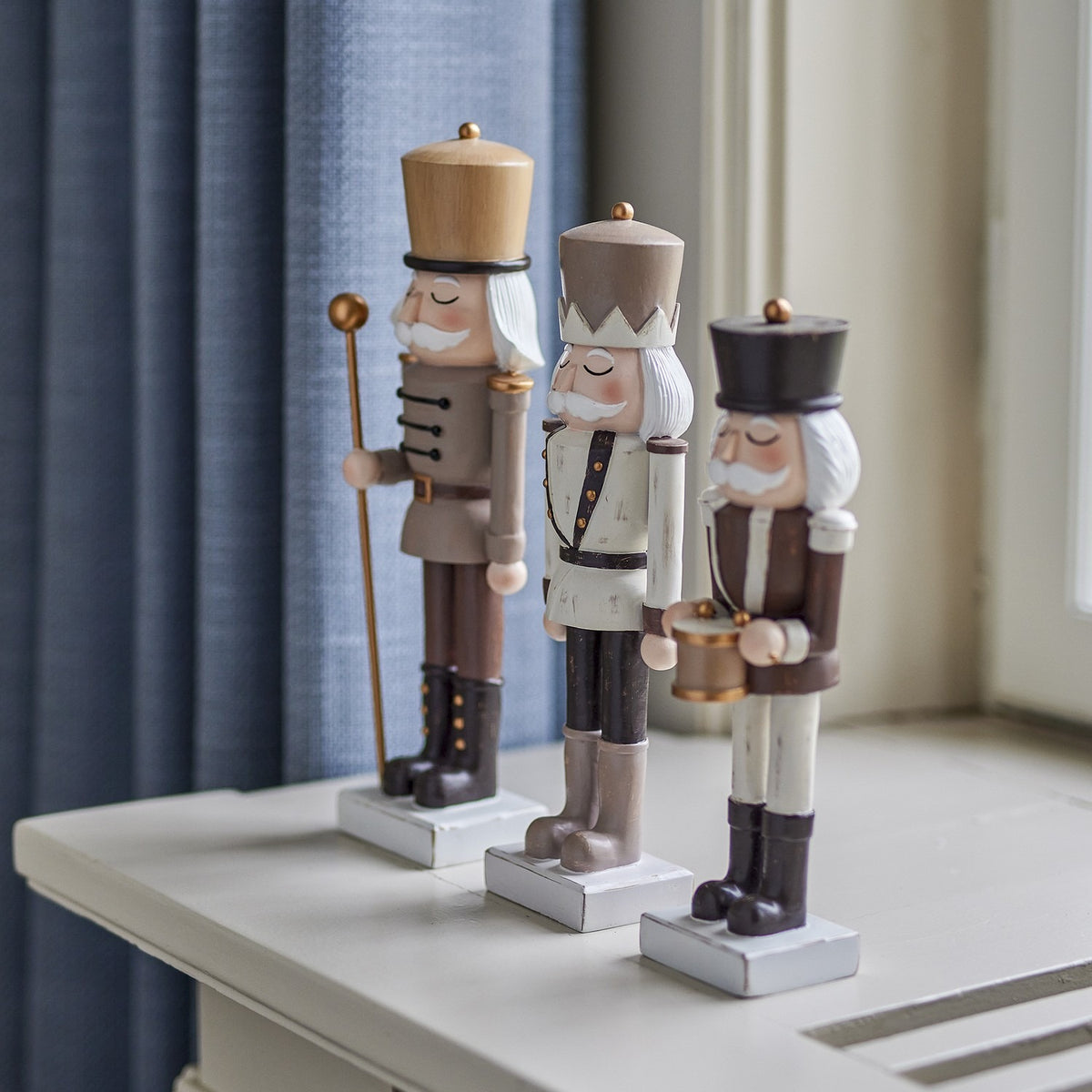Set of three brown-toned resin nutcracker figures with tall hats, moustaches, buttoned jackets, and staff, drum, and crown details, displayed by a window.