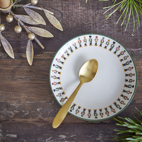 Jolly Stoneware Christmas Plate in cream, 16cm side plate with a green-edged rim featuring a row of nutcracker figures, shown with a gold spoon on a dark wood table.