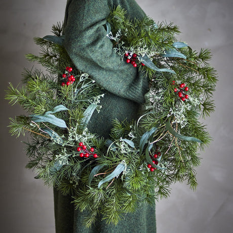 Pine Artificial Christmas Wreath 60cm in green, round and dense with slender leaves, feathery sprigs and red berries, draped over a green knit garment.