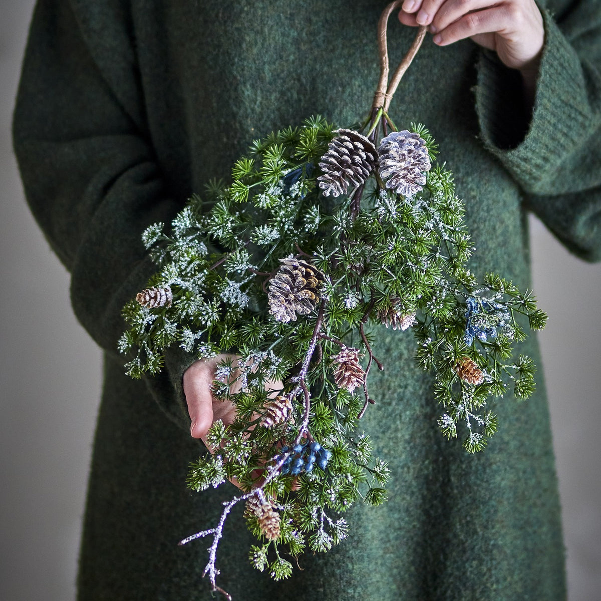 Cedar Christmas Decoration in green, a hanging faux spray with frosted needle tips, pinecones, and berries, held against a green jumper.