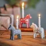 Siffer Horse Resin Christmas Candle Holders, set of 3 folk-style horses in grey, red and white with hand-painted hearts, snowflakes and bird motifs, each holding a lit taper candle on a wooden table.