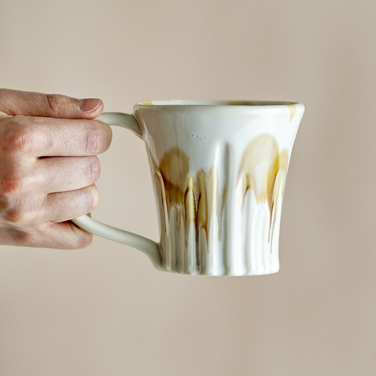 Hand holding a Joseph stoneware mug with ribbed sides and a cream base, amber drip glaze running down, rounded loop handle, against a plain beige background.