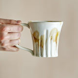 Hand holding a Joseph stoneware mug with ribbed sides and a cream base, amber drip glaze running down, rounded loop handle, against a plain beige background.