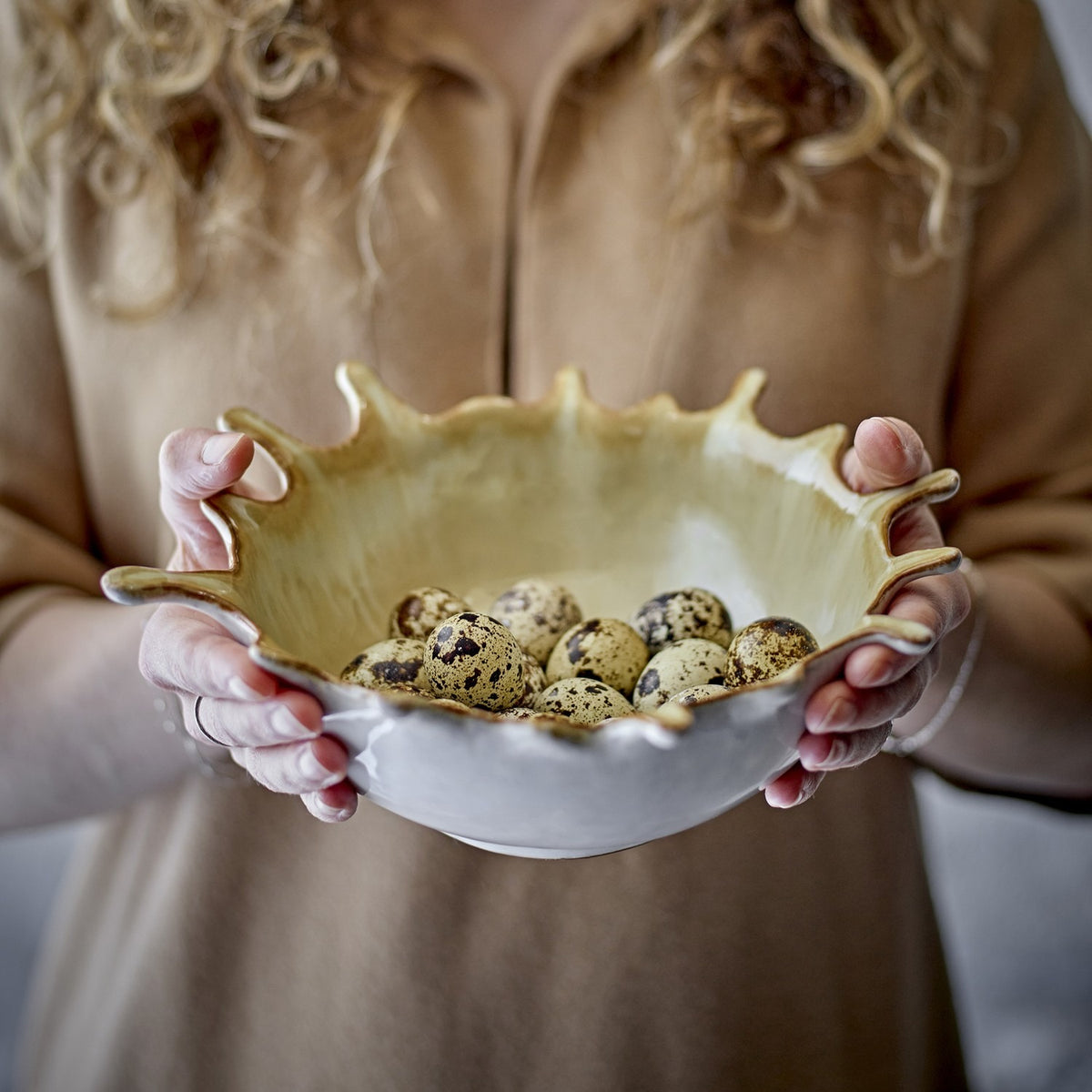 Alcott Stoneware Bowl 24x21cm in beige with a wavy, splash-edged rim, held in two hands and filled with speckled quail eggs.