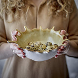 Alcott Stoneware Bowl 24x21cm in beige with a wavy, splash-edged rim, held in two hands and filled with speckled quail eggs.