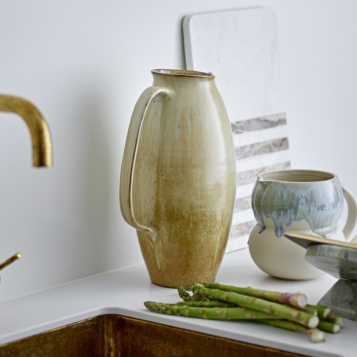 Saskia Speckled Stoneware Vase 31.5cm in beige, jug-shaped with a looped handle, shown on a kitchen counter beside asparagus and ceramic mugs.