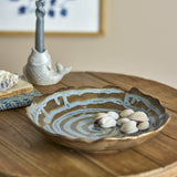 Marjorie Round Stoneware Tray in brown with scalloped rim and spiral glaze pattern, shown with shells on a wooden table in a living room setting.