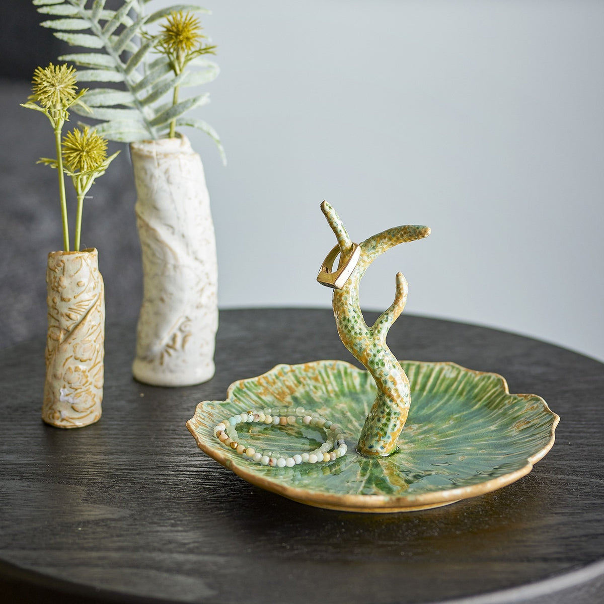 Litton Round Stoneware Tray in green, a leaf-shaped ceramic dish with ribbed, scalloped edges and a branch-like ring holder, shown with a beaded bracelet beside textured vases.