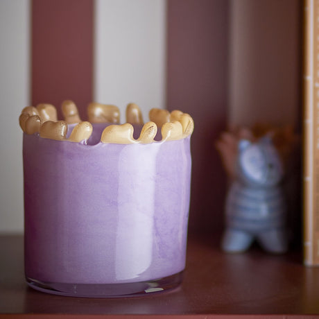 Edger Wavy Glass Tealight Holder in purple, a round glass vessel with a mottled cloudy finish and a wavy rim of rounded gold-toned glass drops, shown on a shelf.