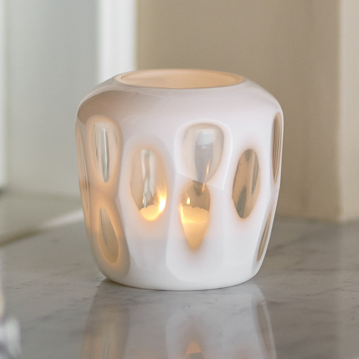 Rohan Glass Tealight Holder in white, a rounded glass votive with recessed teardrop cutouts glowing from a lit candle inside, shown on a marble surface.
