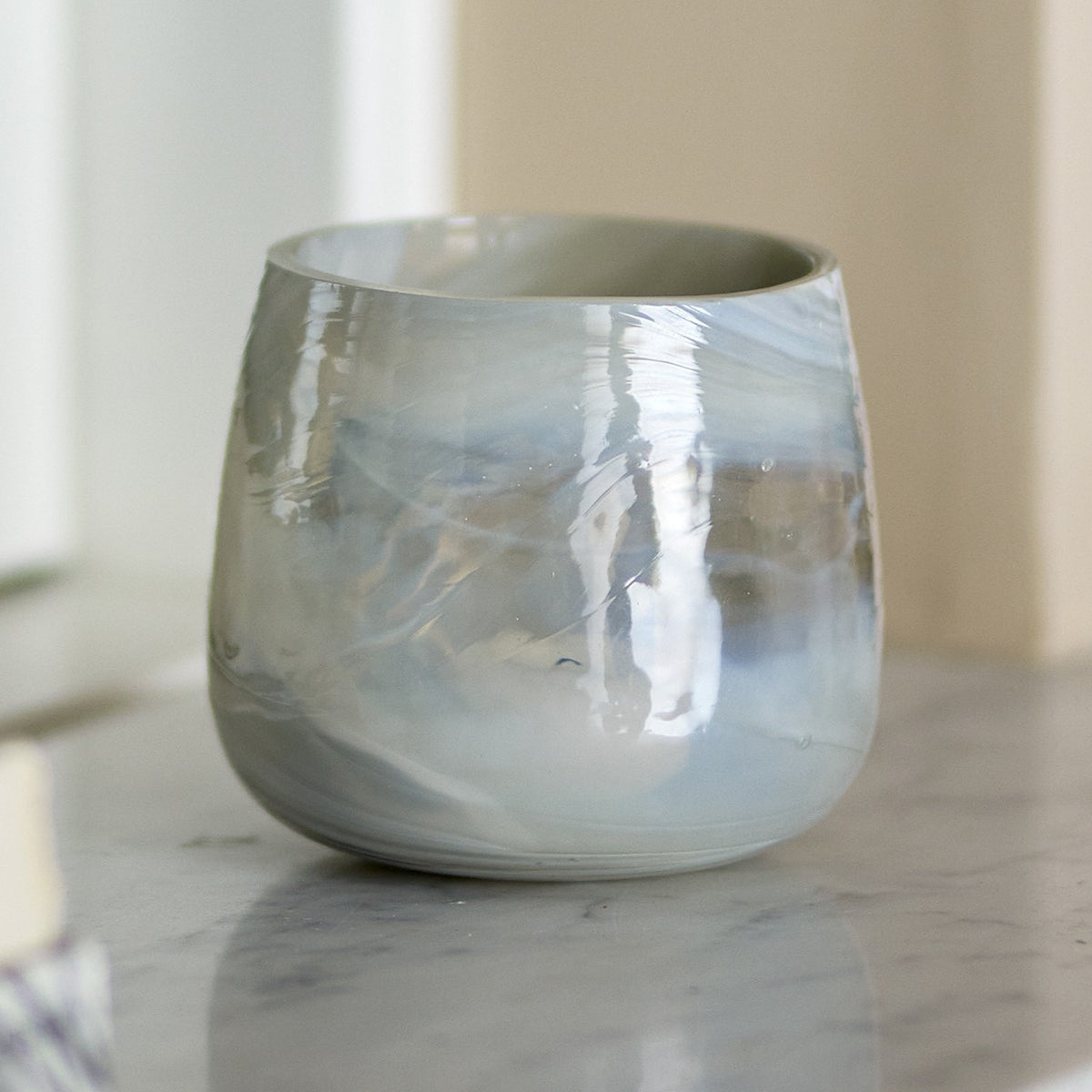 Imran Marbled Glass Tealight Holder in grey, a rounded tub-shaped glass votive with swirled marbling, shown on a marble surface in a living room setting.