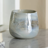 Imran Marbled Glass Tealight Holder in grey, a rounded tub-shaped glass votive with swirled marbling, shown on a marble surface in a living room setting.