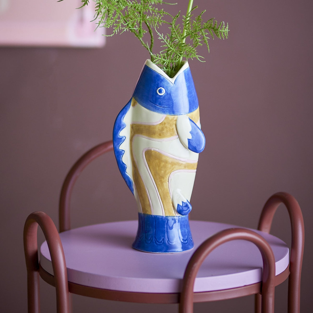 Irving Blue Fish Stoneware Vase, 22cm, shaped like a fish with painted eye and fins, in blue, white and ochre, holding greenery on a chair-side table.