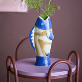 Irving Blue Fish Stoneware Vase, 22cm, shaped like a fish with painted eye and fins, in blue, white and ochre, holding greenery on a chair-side table.