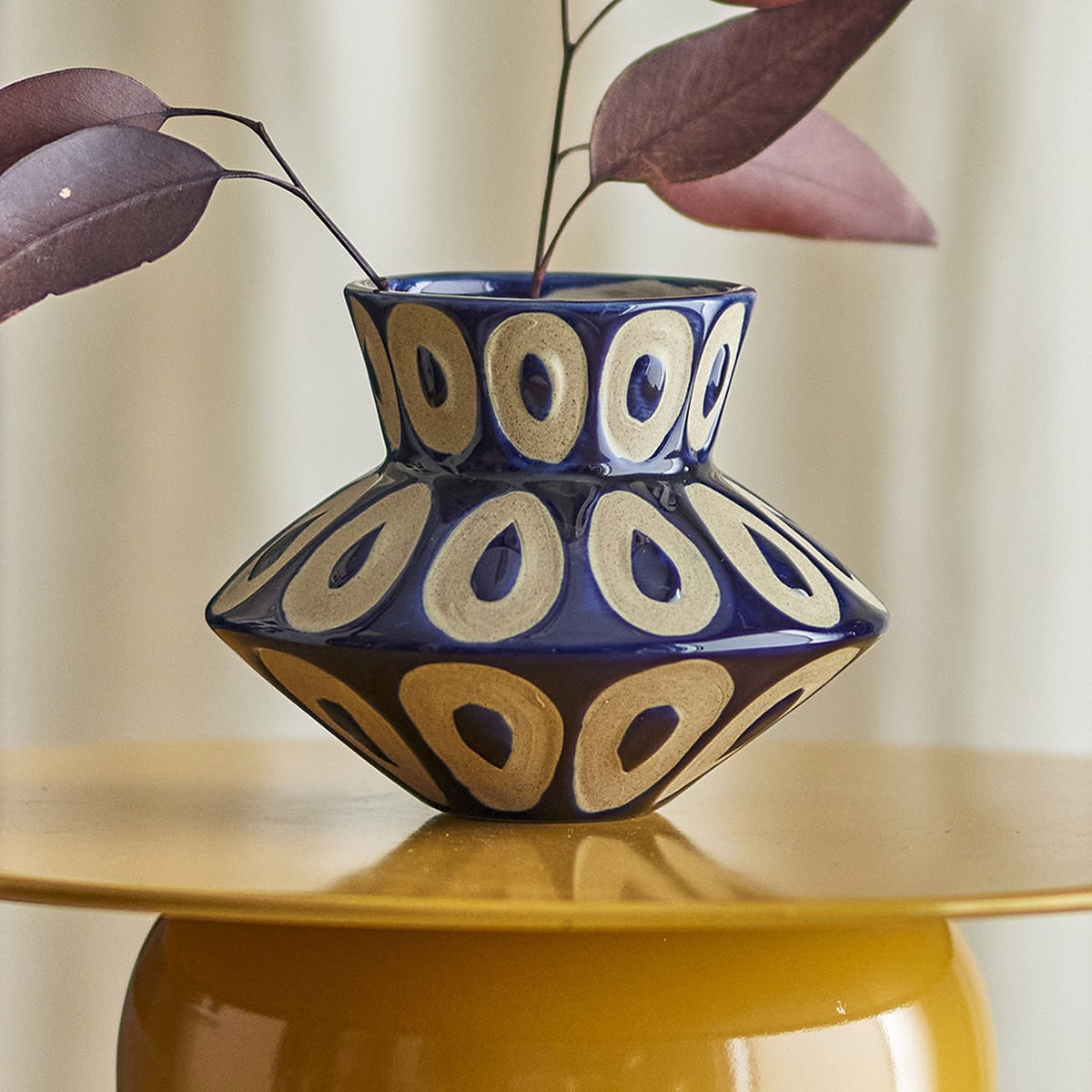 Bashir Stoneware Vase 12cm in blue with a spinning-top shape and speckled ring pattern, holding dried leaves on a yellow side table.