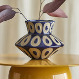 Bashir Stoneware Vase 12cm in blue with a spinning-top shape and speckled ring pattern, holding dried leaves on a yellow side table.