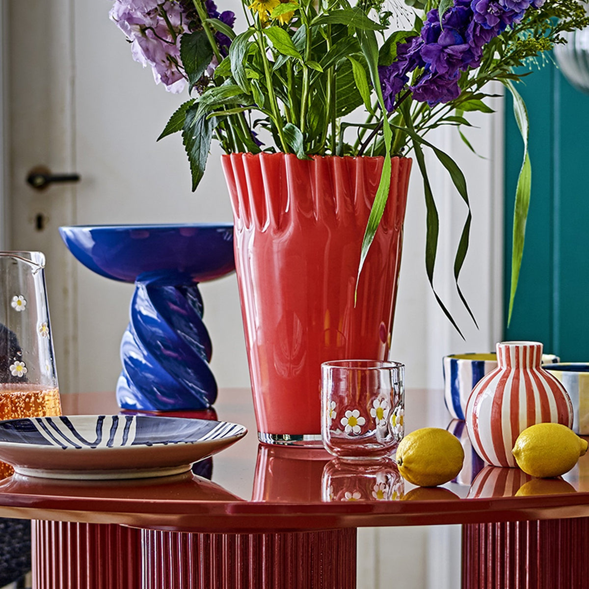 Janine Fluted Glass Vase 30cm in pink with wavy, frilled rim, styled on a red table with purple flowers, lemons, and patterned tableware.