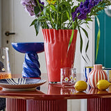 Janine Fluted Glass Vase 30cm in pink with wavy, frilled rim, styled on a red table with purple flowers, lemons, and patterned tableware.