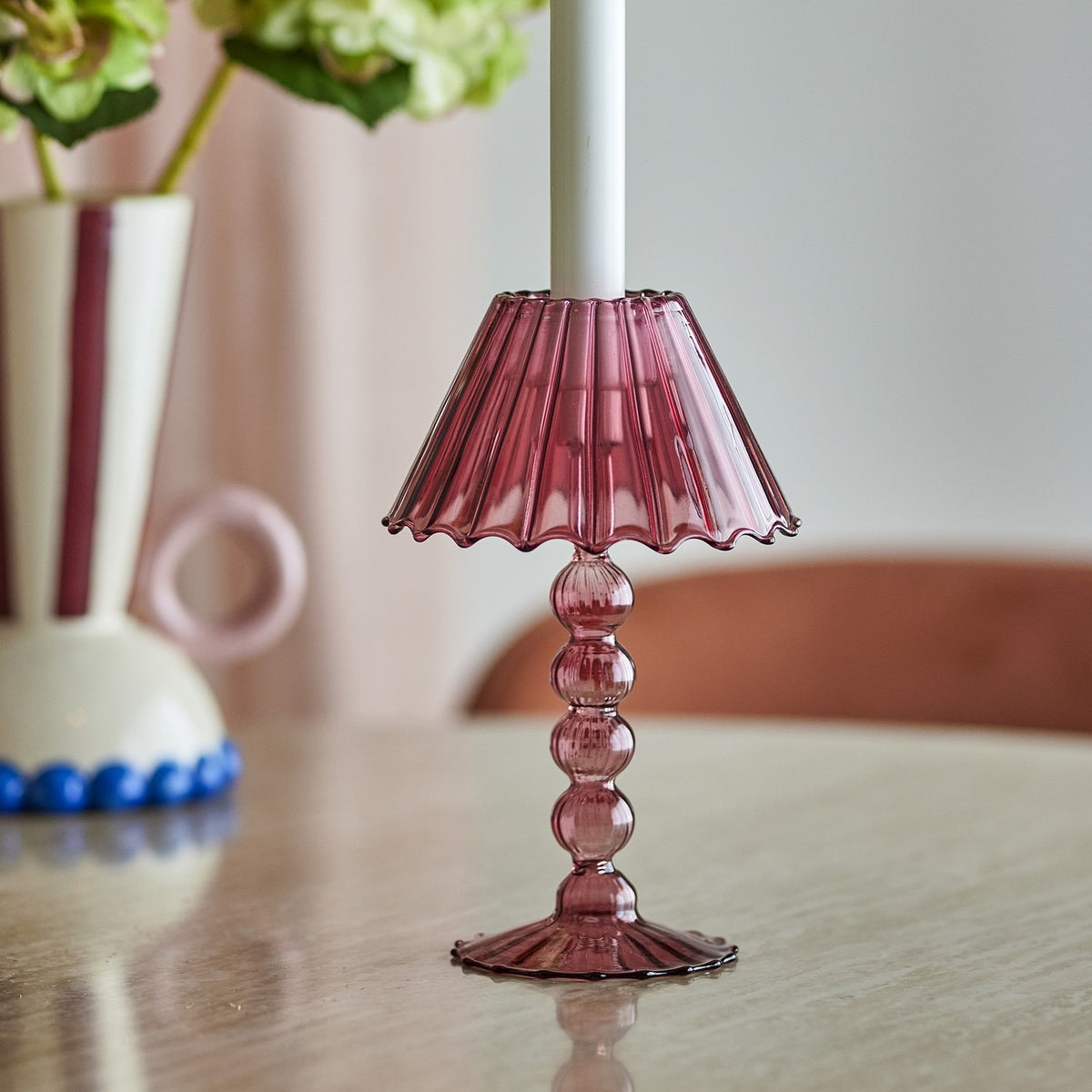 Cisco Glass Tealight Holder 19.5cm in pink, lamp-shaped with a fluted wavy shade and stacked bead stem on a ribbed scalloped foot, shown on a table beside a vase of flowers.