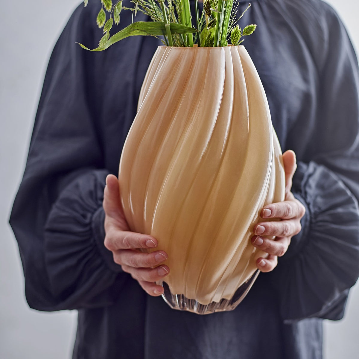 Kanji Glass Vase 30cm in beige, spiral-ribbed glass body with a bud-like curved shape, held in two hands with green stems and foliage arranged inside.