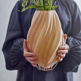Kanji Glass Vase 30cm in beige, spiral-ribbed glass body with a bud-like curved shape, held in two hands with green stems and foliage arranged inside.