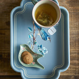 Meggie Tray in blue MDF with scalloped rectangular rim and bird-on-branch illustration, shown holding a cup of tea and biscuits on a wooden table.