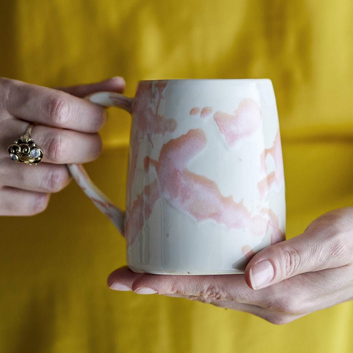 Winnie Marbled Stoneware Mug in white with pink reactive glaze pooling across the rounded body, held by hand via its open loop handle against a yellow backdrop.