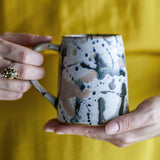 Hands holding a Winnie stoneware mug with a multicoloured reactive splatter glaze, dripped speckled pattern and matching handle, against a mustard yellow background.