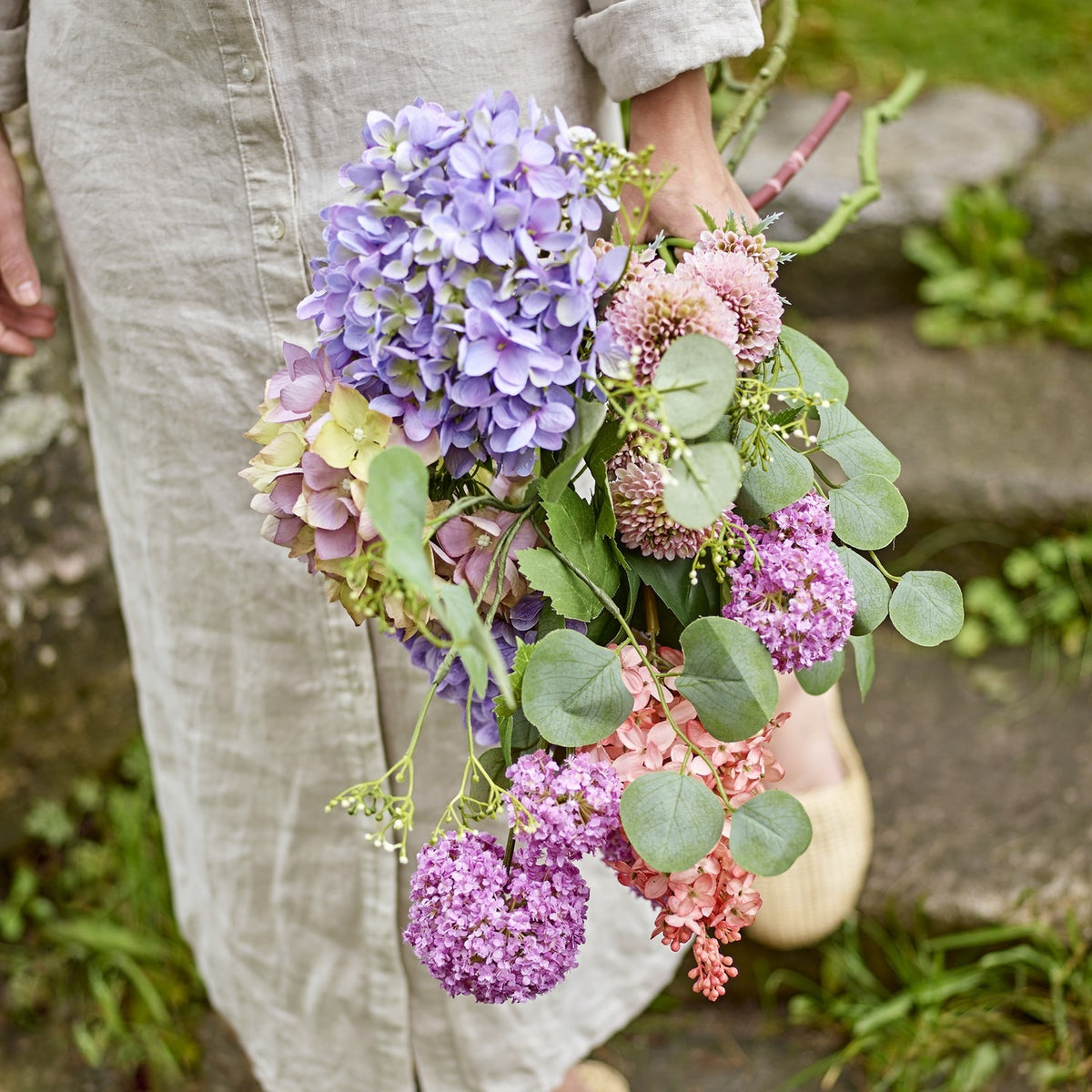 Edge Artificial Bouquet in Purple, held outdoors, showing hydrangea-style clusters, pompom heads, and trailing eucalyptus-style leaves in purple and pink tones.