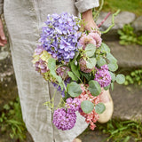 Edge Artificial Bouquet in Purple, held outdoors, showing hydrangea-style clusters, pompom heads, and trailing eucalyptus-style leaves in purple and pink tones.