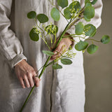 Eucalyptus Artificial Stem 82cm in Green held in hand, showing rounded leaves and fine bud clusters along slender branches against a neutral linen backdrop.