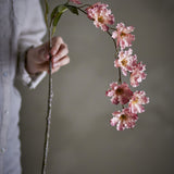 Wild Cherry Artificial Stem in pink, held by hand, showing a knotted stem arching into ruffled fabric blossoms and leaves against a neutral background.