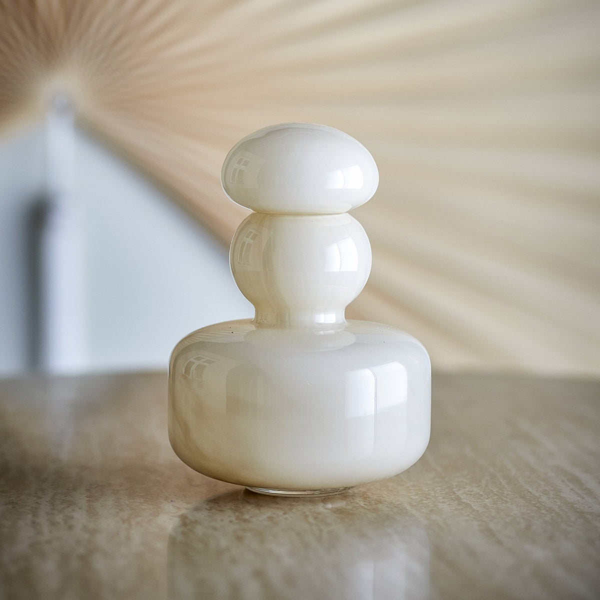 Cynthia Glass Vase 21cm in Beige, a glossy mouth-blown glass vase with a stacked design of a broad base, ball-shaped neck and mushroom-domed stopper, shown on a reflective tabletop.