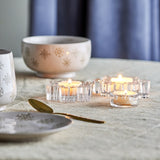 Dania Snowflake Glass Christmas Tealight Holders, set of 3 clear glass snowflake-shaped candle holders with lit tealights, styled on a table with bowls and a spoon.