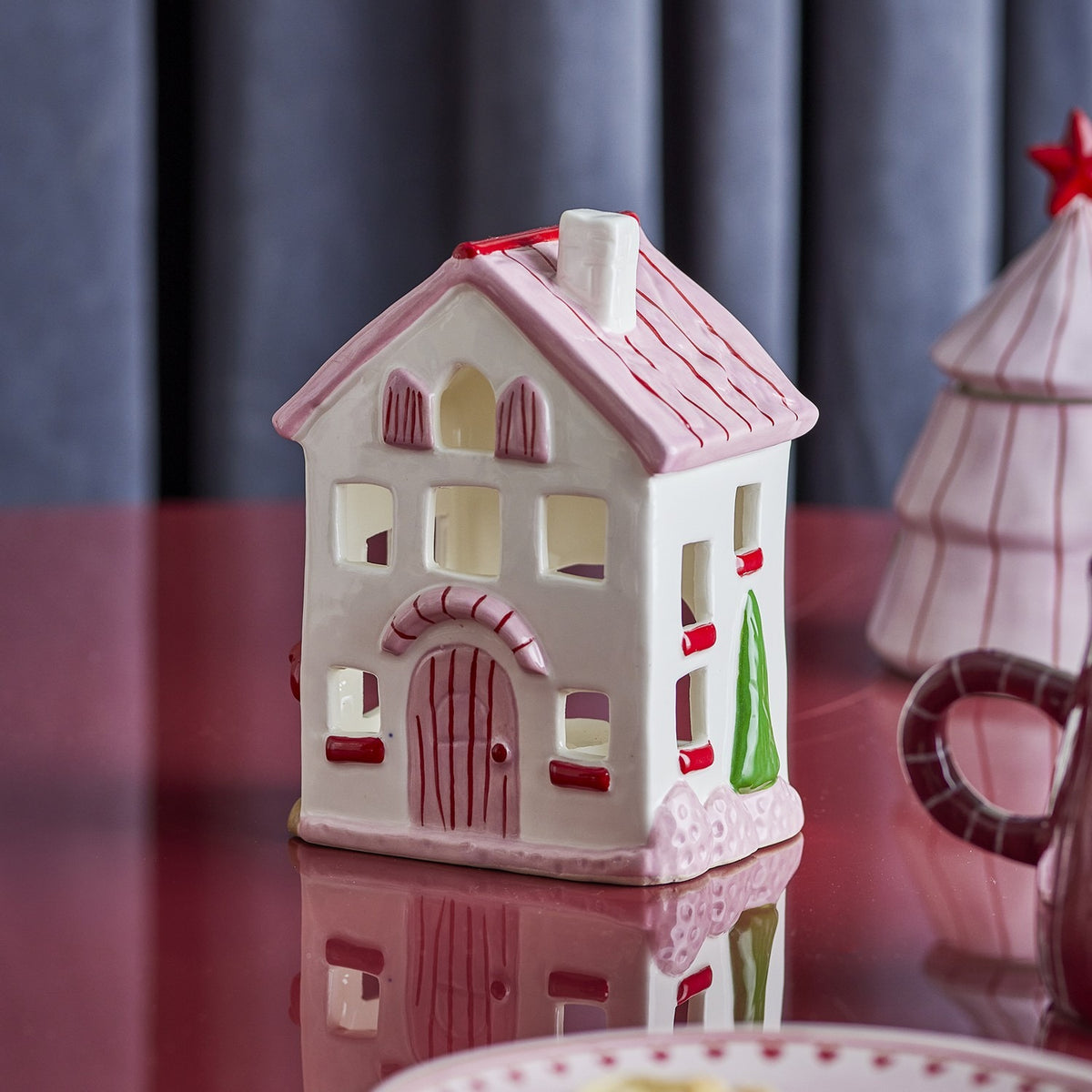 Shirly House stoneware Christmas tealight holder in white, with pink roof and base, red door and windows, and green tree detail, shown on a dark red table.