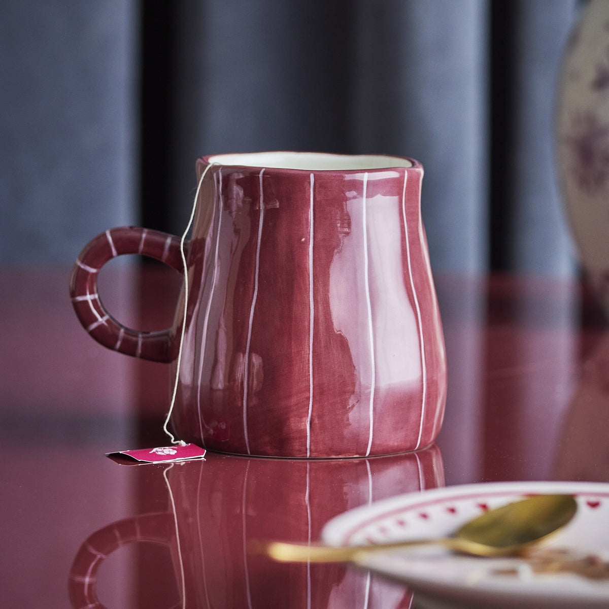 Burgundy stoneware mug from the Shirly striped set, hand-painted with fine vertical white stripes, glossy finish, ring handle, shown on a glass surface with a tea bag.