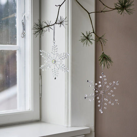 Miku Snowflake Christmas Tree Decorations, Set of 2, clear beaded wire snowflakes hanging from a pine branch beside a white-framed window.