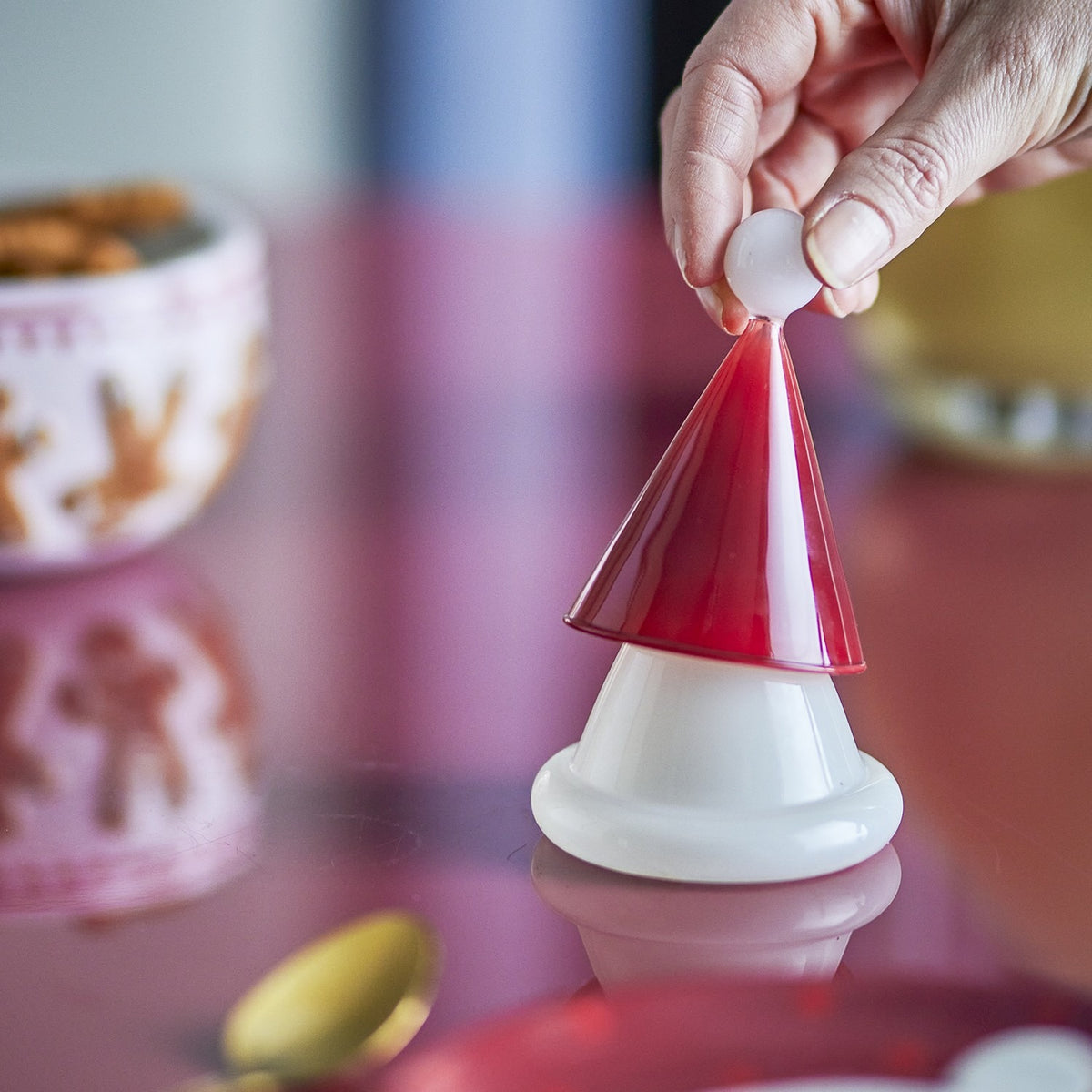Patrice Glass Christmas Toothpick Holder in red, shaped like a Father Christmas hat with a white bobble tip, held by hand above a glossy white base on a reflective tabletop.