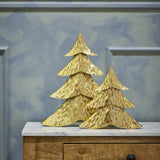 Kaiam Iron Christmas Decorations Set of 2 in gold, tiered fir-tree shapes with rippled textured surfaces, displayed on a marble-top wooden cabinet against a pale blue panelled wall.