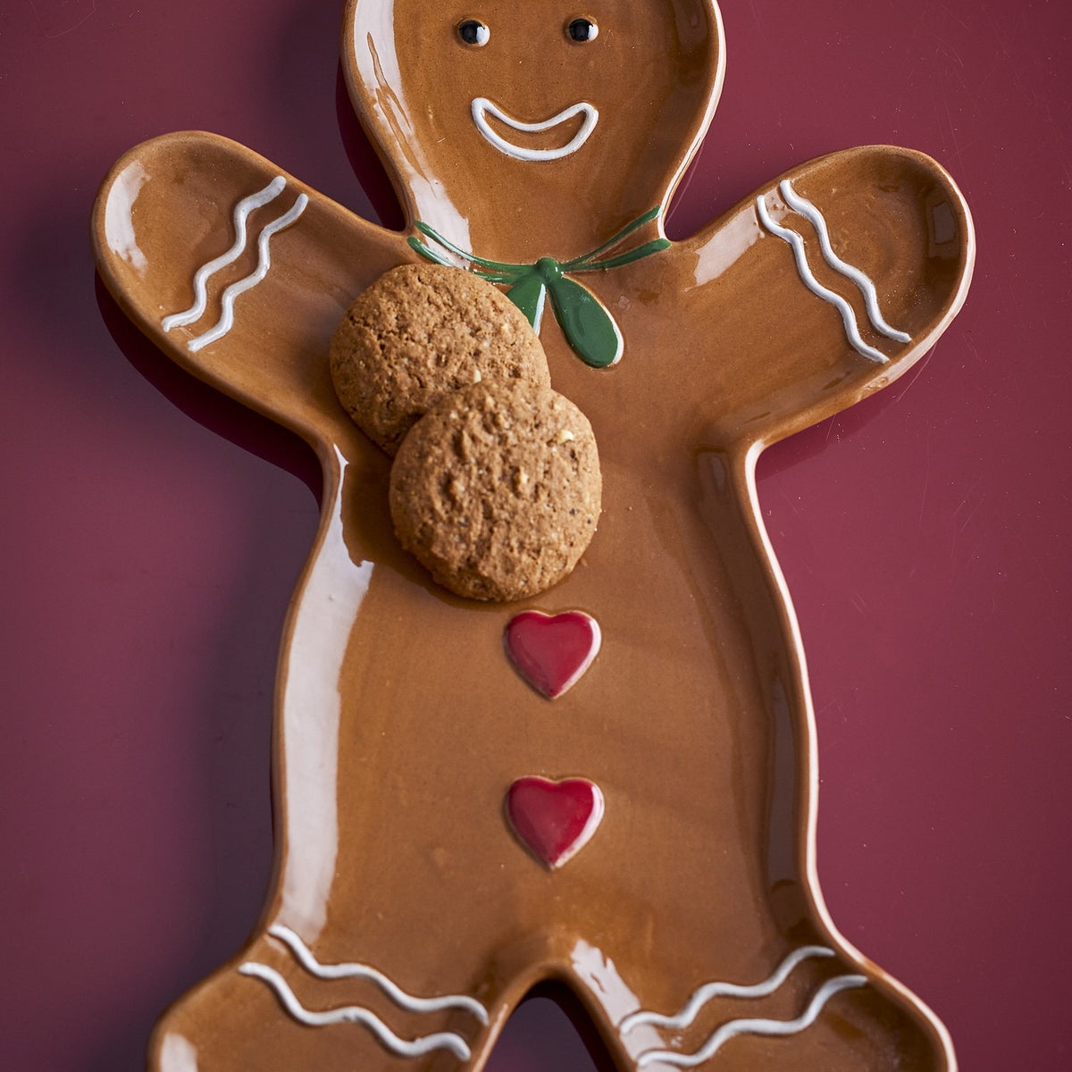 Ginnie Gingerbread Man Stoneware Christmas Platter in glossy brown, with smiling face, green bow, red hearts and icing details, shown holding two cookies on a dark red background.