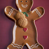 Ginnie Gingerbread Man Stoneware Christmas Platter in glossy brown, with smiling face, green bow, red hearts and icing details, shown holding two cookies on a dark red background.