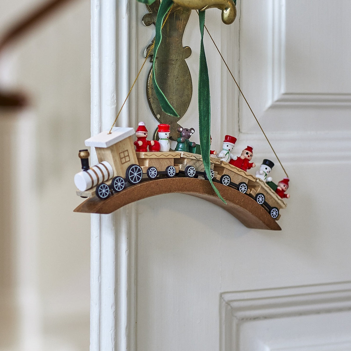 Bertie Wooden Christmas Tree Decoration in beige birch wood, shaped as a steam train with painted passenger figures, hanging from a green ribbon on a white door.