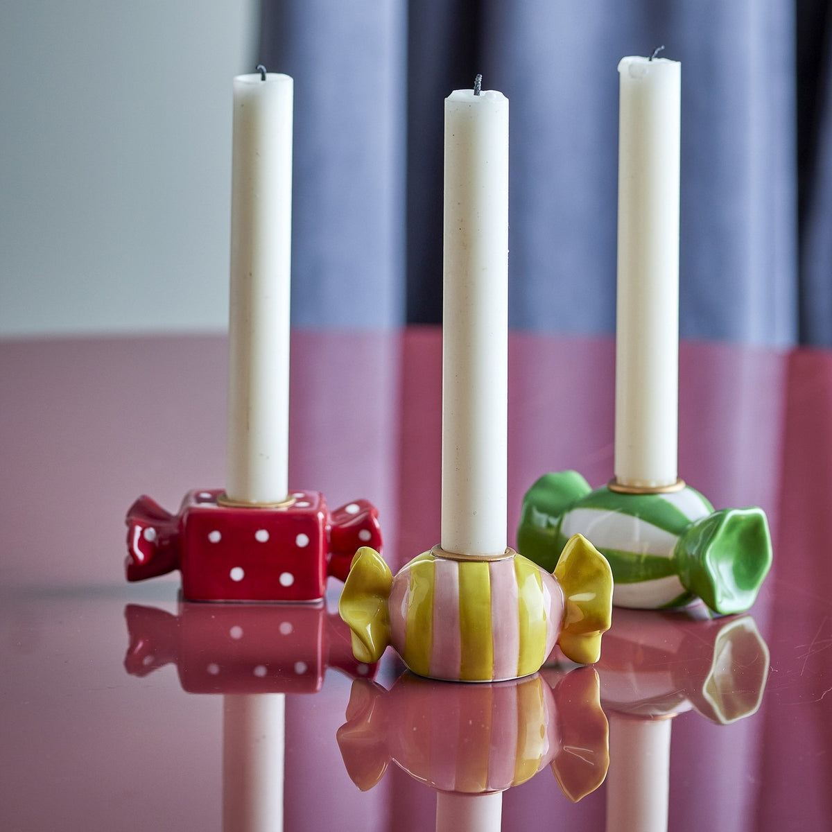 Set of three wrapped-sweet stoneware candle holders in red polka-dot, yellow-striped, and green designs, each holding a white taper candle on a glossy pink surface.