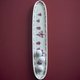 Cream stoneware Christmas platter with a long oval shape, hand-painted pine tree motif, and branch-patterned rim, shown with three white truffles on dark red.