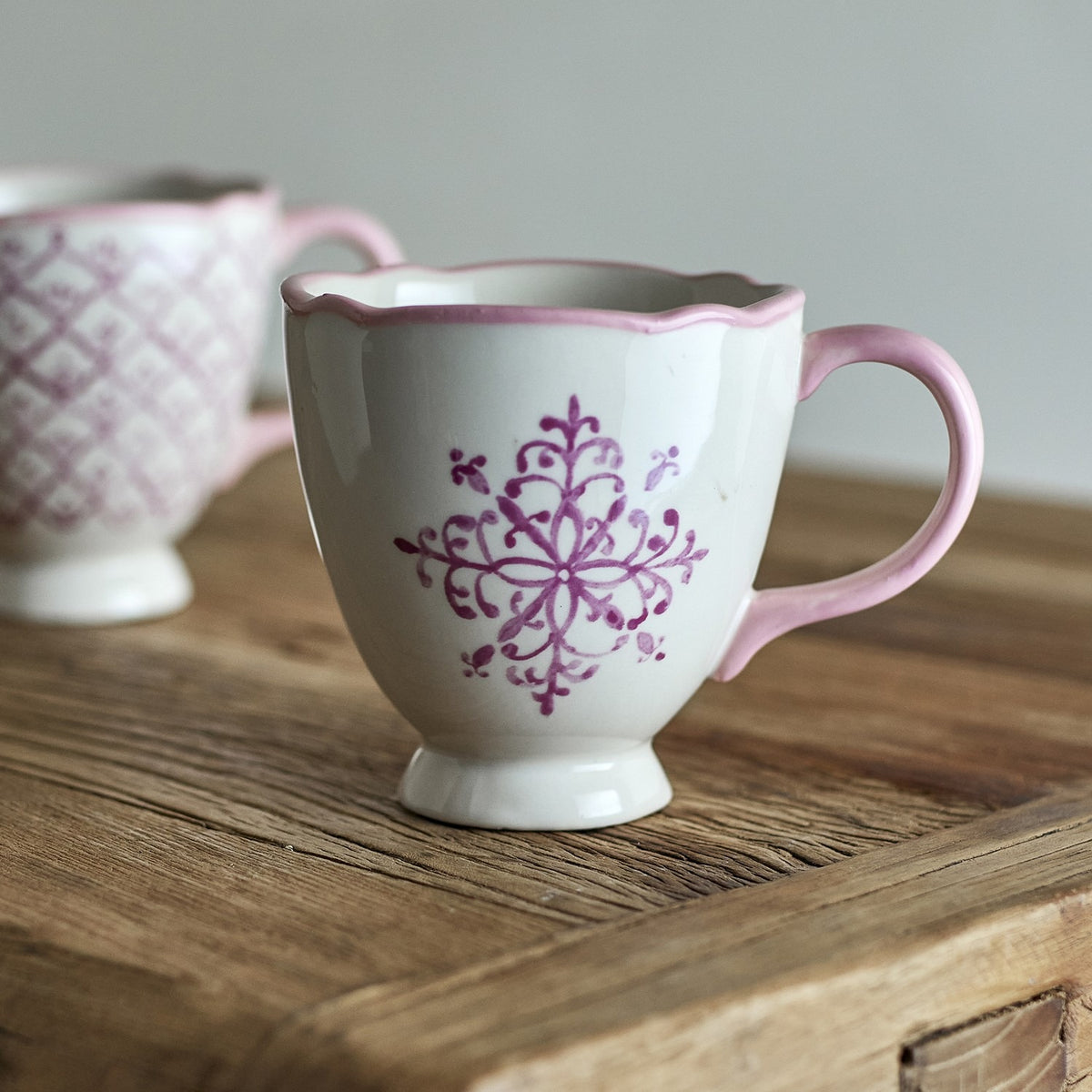 White stoneware Christmas mug from the Wonderland set with a pink scalloped rim and handle, footed base, and hand-painted purple snowflake motif on a wooden surface.