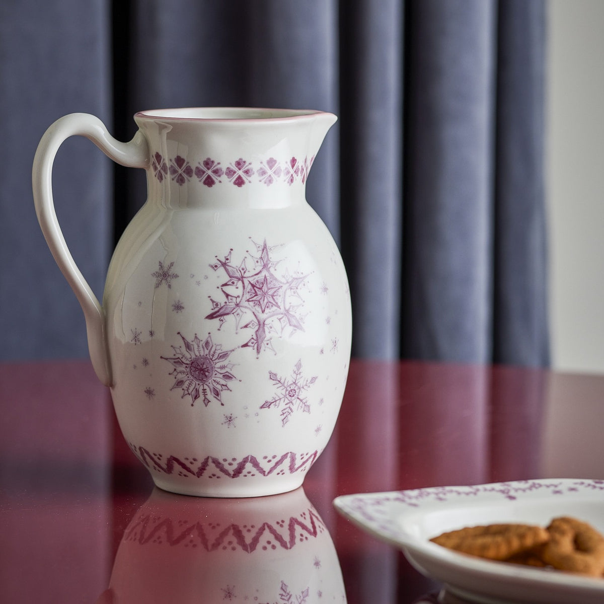 White stoneware Christmas jug with looped handle and pouring lip, decorated with purple snowflake motifs, displayed on a dark red table with a biscuit plate nearby.