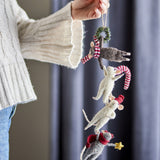 Hanging wool Christmas decoration held by hand, showing four felt mice in red-and-white hats linked in a chain below a small wreath, against a curtained room backdrop.